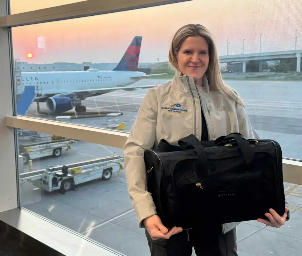 Monica from Cabin Companions, a professional cat flight nanny, stands in front of a Delta plane at sunrise holding a soft-sided pet carrier for in-cabin pet travel.